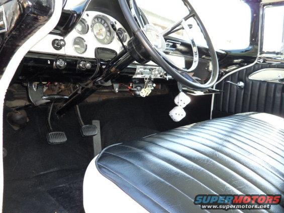 Interior shot of a classic car showing the dashboard, steering wheel, and front seat. Dice hang from the rearview mirror