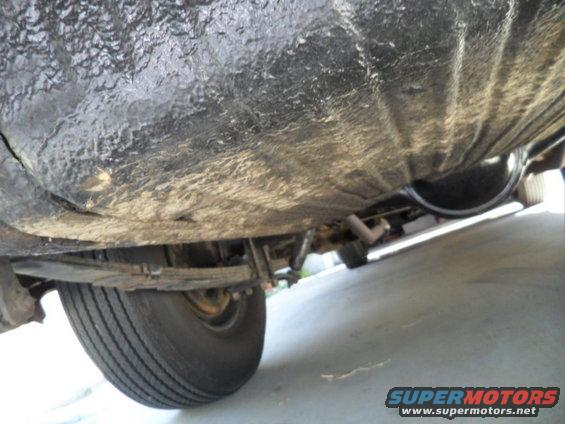 Underside view of a car, showing the frame, exhaust pipe, and rear tire. The surface is coated in black undercoating