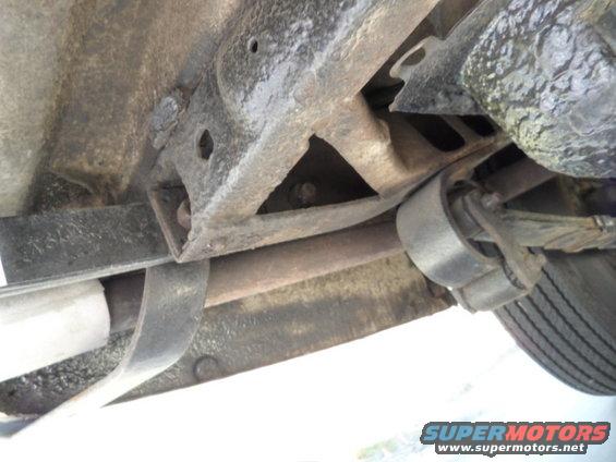 Undercarriage of a vehicle showing rusty frame, leaf springs, and exhaust pipe. A tire is partially visible