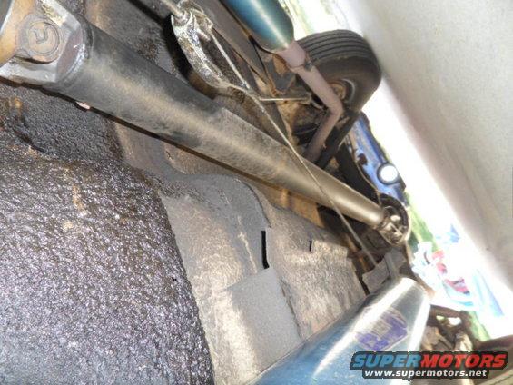 Underside view of a car's drivetrain and undercarriage. Shows the driveshaft, exhaust pipe, and a tire