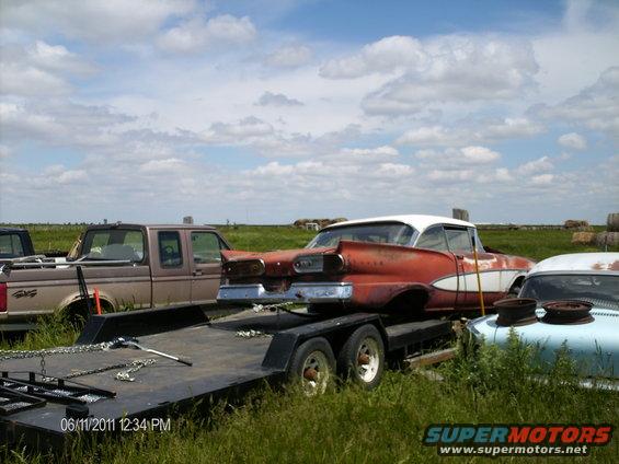Red and white 1958 Ford Fairlane on trailer next to a pickup truck and a light blue car