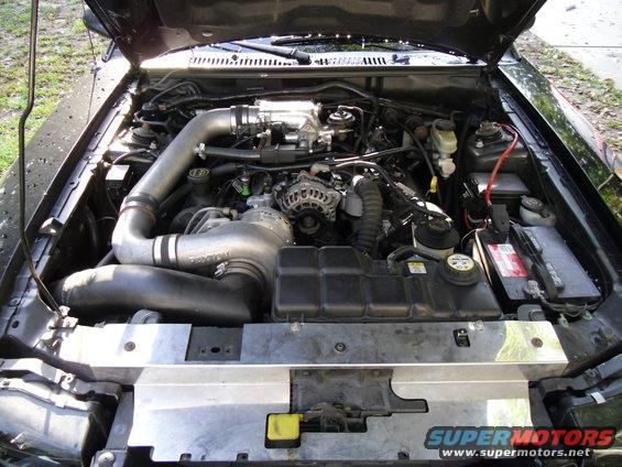 Black engine bay with exposed engine, intake, battery, and radiator hoses. Supermotors.net logo visible