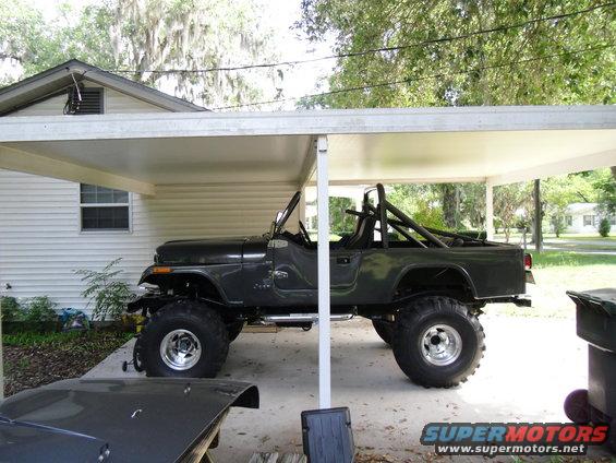 Dark green Jeep CJ-7 with roll cage and large tires parked under a carport