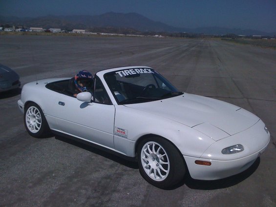 White convertible sports car with a driver wearing a helmet, parked on a tarmac with mountains in the background. Has a 'Tire Rack' banner on the windshield