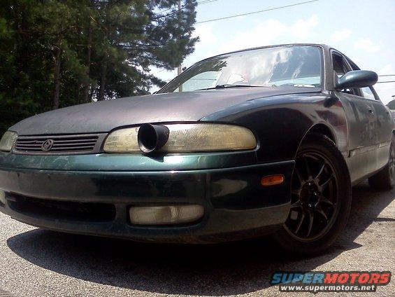 Green 1994 Mazda 626 with custom exhaust pipe in headlight, front view