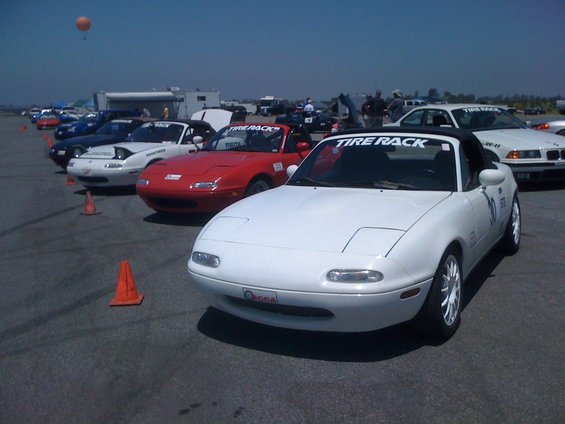 White Mazda Miata with pop-up headlights and a black soft top, parked on asphalt with orange cones