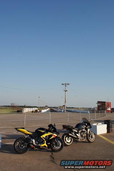 Two sport motorcycles, one yellow and black, one dark gray, parked on asphalt near a drag strip