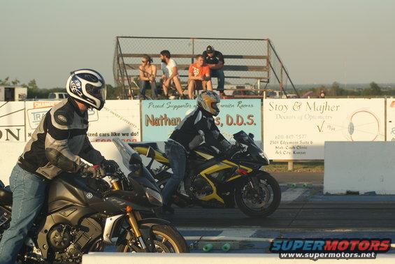 Two motorcycles race on a drag strip with spectators in the background