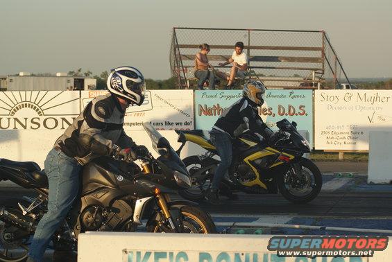 Two motorcycles lined up on a drag strip, riders in gear, spectators in bleachers behind them