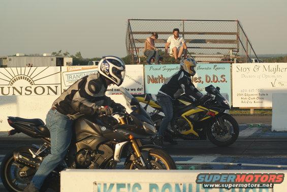 Two motorcycles racing on a drag strip, with spectators in the background