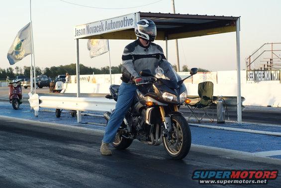 Motorcycle on drag strip, rider in gear. Scooter in background. Sign says Jason Novacek Automotive