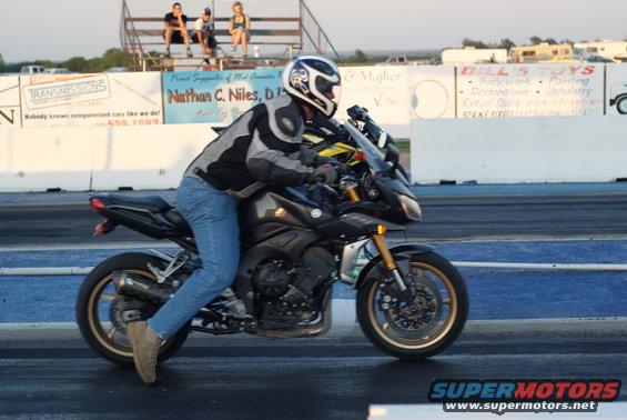 Motorcyclist in full gear on a black sportbike at a drag strip with spectators in the background