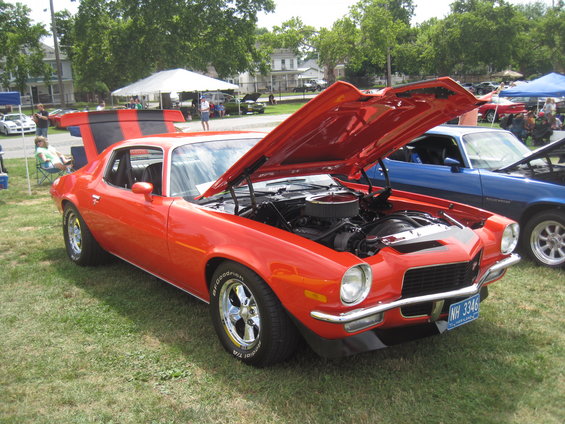 orange muscle car with hood open, black stripes on spoiler, parked on grass next to blue car