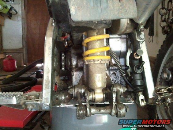 Motorcycle rear suspension with yellow spring shock absorber, silver frame, and foot peg. A red gas can is visible in the background