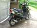 2008 Yamaha Zuma - A black scooter is parked on a concrete surface next to a large storage unit, with greenery in the background.