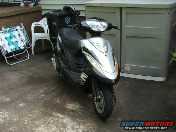 Black and silver scooter parked on concrete, front view. A black storage bin is behind it