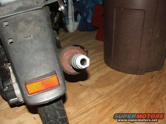 Rear of a dark-colored scooter showing a textured fender, orange reflector, and a rusty exhaust pipe with a white nut