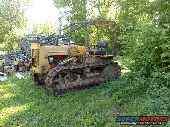 Old yellow crawler tractor with tracks sits in grass next to a pile of logs. It has a roll cage and a single seat