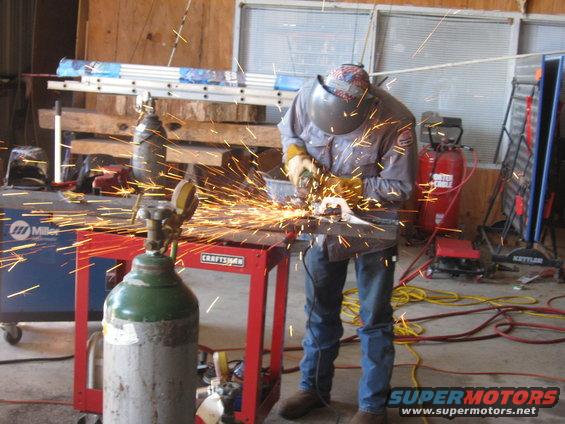 Man in welding mask and gloves grinding metal, creating many sparks. A welding tank and red Craftsman cart are visible