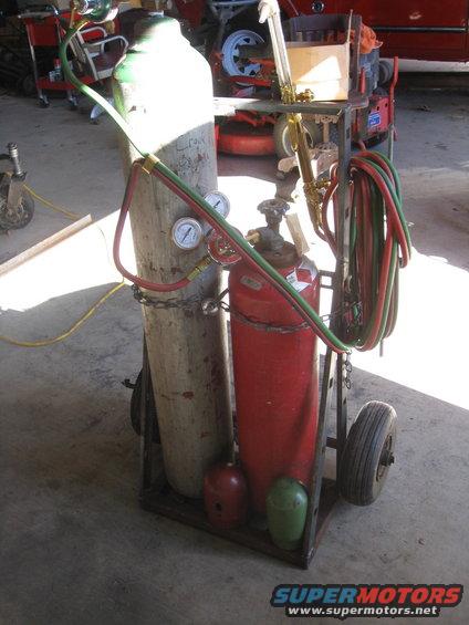 Welding cart with two gas tanks, hoses, and a torch. Some lawn equipment is in the background