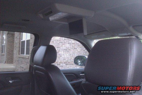 Interior view of a vehicle showing leather seats, a ceiling-mounted screen, and a brick building visible through the window