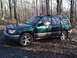 1999 Subaru Forester - A green Subaru Forester is parked on a dirt road surrounded by trees.