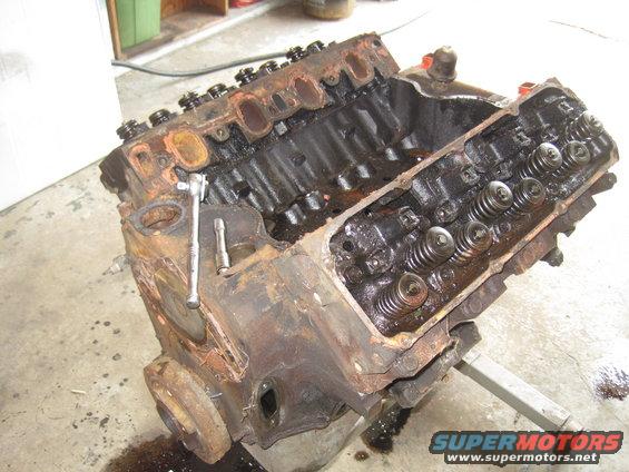 Rusty V8 engine block with valve covers removed, showing springs and pushrods. A ratchet is on the block
