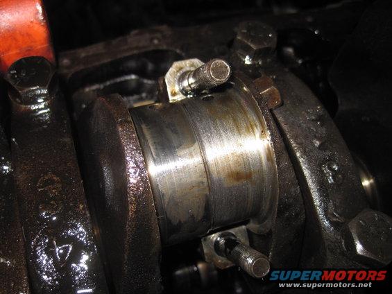 Engine crankshaft bearing with oil residue and visible wear marks. A bolt is partially threaded into the bearing cap