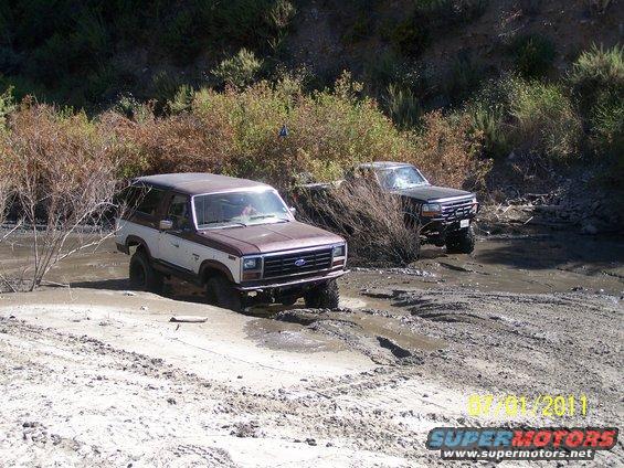 Brown and white 1980s Ford Bronco stuck in mud, front view. Another black truck behind it