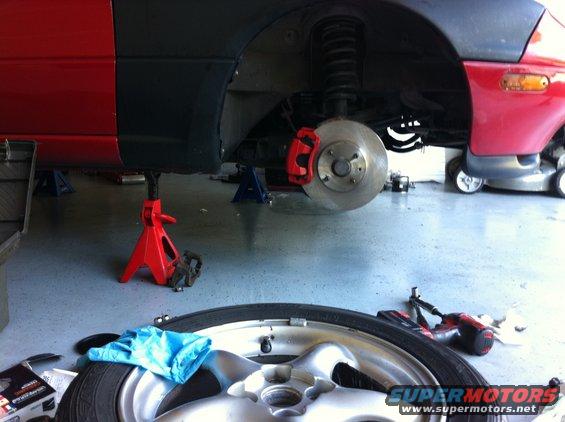 Car on jack stands with front wheel removed, showing brake rotor and caliper. Tools and a spare tire are on the floor