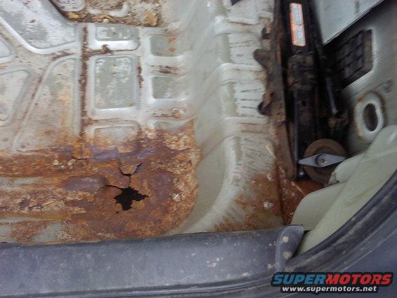 rusted out floor pan of a car, showing a hole and some jack equipment