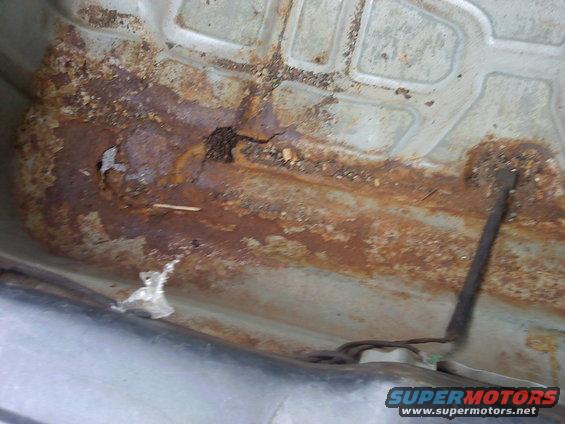 Undercarriage of a vehicle with significant rust and corrosion, showing a cracked metal panel and exposed wires
