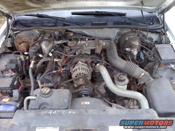 Engine bay with V8 engine, hoses, and belts visible. Some writing on the radiator cover