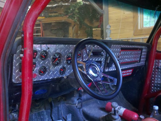 Interior of a red off-road vehicle with a diamond plate dashboard, multiple gauges, and a racing steering wheel