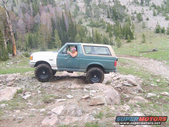 1989 Ford Bronco driving over rocks on a mountain trail, white front fender, green body