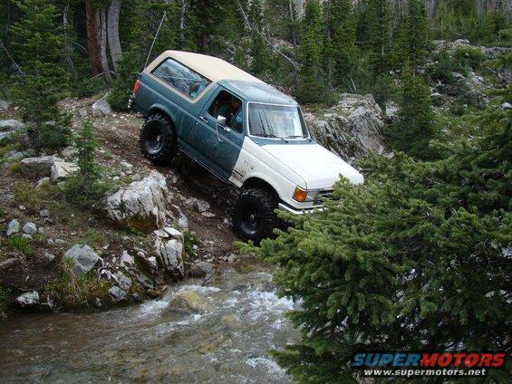 Two-tone green and white Ford Bronco off-roading through a stream in a forest