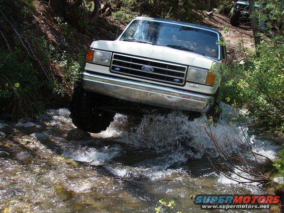 White 1987 Ford Bronco crossing a stream, with another vehicle visible in the background