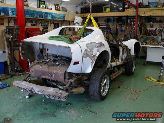 White car body, missing engine and interior, suspended by yellow straps. Rust and peeling paint visible on rear quarter panel