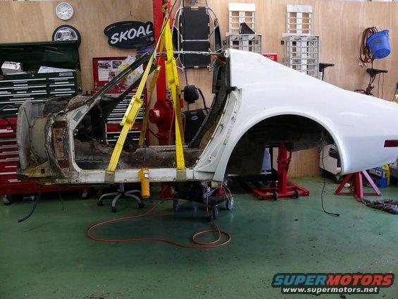 White car body on repair stands, lifted by yellow straps, showing disassembled interior and rear quarter panel
