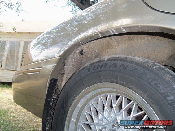 Close-up of a car's rear wheel well, showing the tire and part of the fender. A house is visible in the background