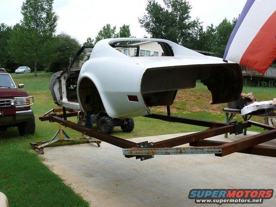White Corvette body on a trailer, with a red pickup truck and a silver car in the background