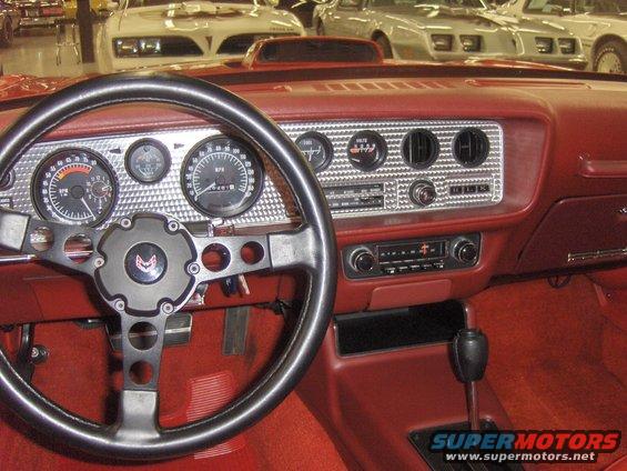 1977 Pontiac Firebird interior, red dashboard with gauges, steering wheel, and shifter. Other cars visible in background