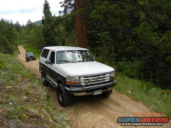 White and teal Ford Bronco SUV on a dirt road, front quarter view. Modified with off-road tires and a brush guard