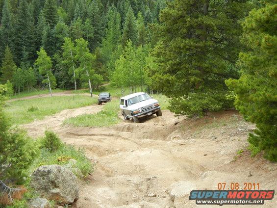 White SUV driving down a steep dirt trail with a dark SUV behind it, surrounded by trees