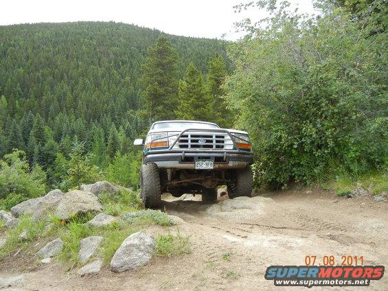 Ford truck with a brush guard driving over rocks on a dirt trail, trees and a hill in the background