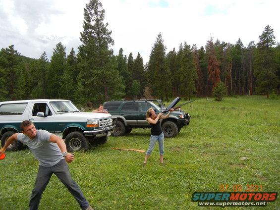 White and teal 1996 Ford Bronco parked in a grassy field with a man throwing a disc