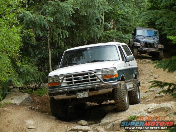 White and green Ford Bronco with a brush guard driving over rocks on a trail, with a Jeep Wrangler behind it