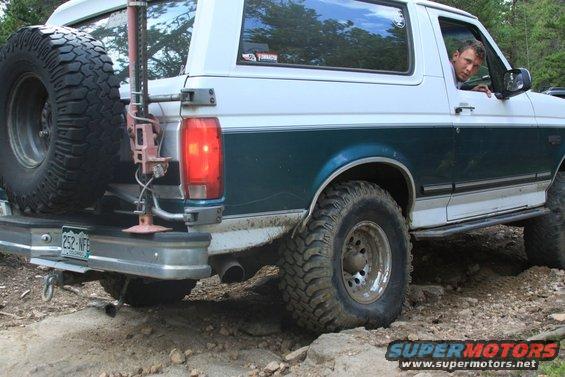White and teal Ford Bronco with spare tire on back, driving over rocks