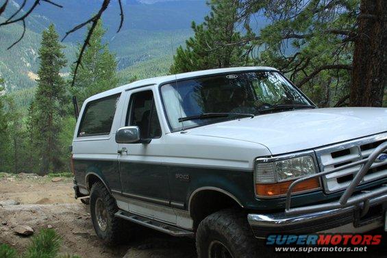 White and green Ford Bronco on a dirt road with trees and mountains in background