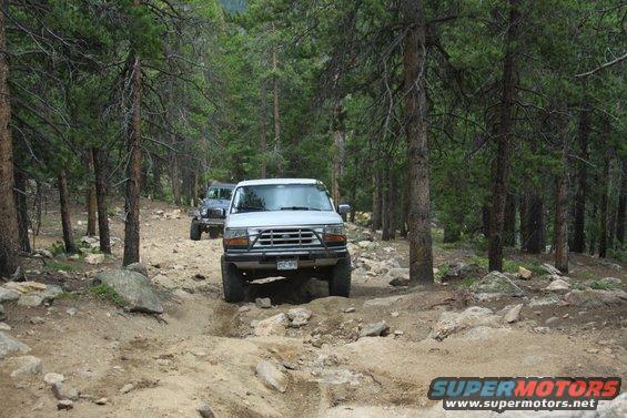 White 1992 Ford Bronco and a grey Jeep on a rocky, wooded trail